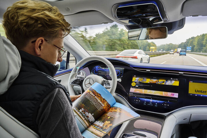 Mann sitzt in Cockpit und testet automatisiertes Fahren. Er lenkt nicht mehr selbst sondern ließt während des Fahrens eine Zeitschrift.