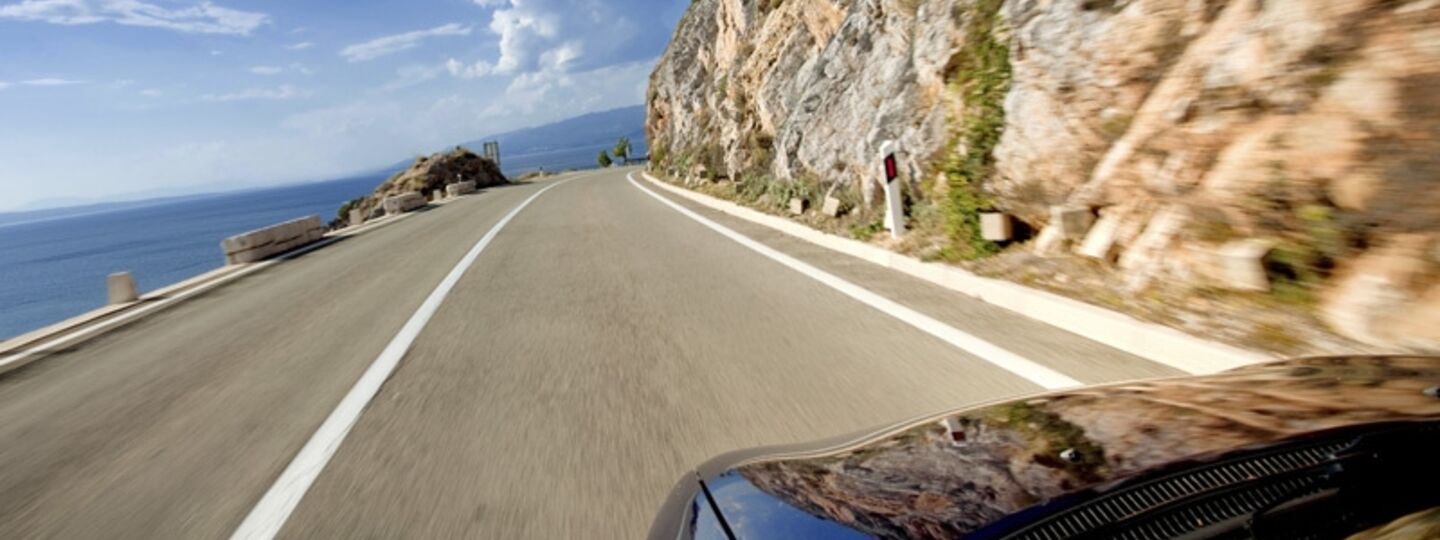 Ein Auto fährt entlang einer Küstenstraße mit einer steilen Felswand auf der rechten Seite.