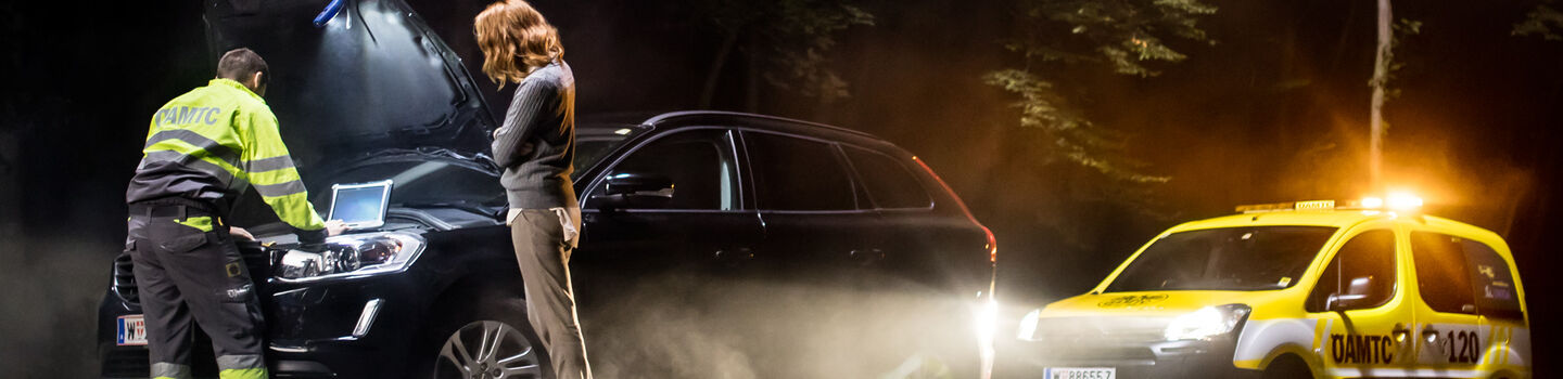 An einer Freilandstraße steht ein dunkler Combi, davor steht eine Frau. Die Motorhaube ist geöffnet und ein ÖAMTC Pannenhelfer schaut sich den Motor an. Es ist Nacht und das ÖAMTC Pannenhelferauto steht mit leuchtenden Scheinwerfern hinter dem schadhaften PKW und leuchtet die Situation aus.