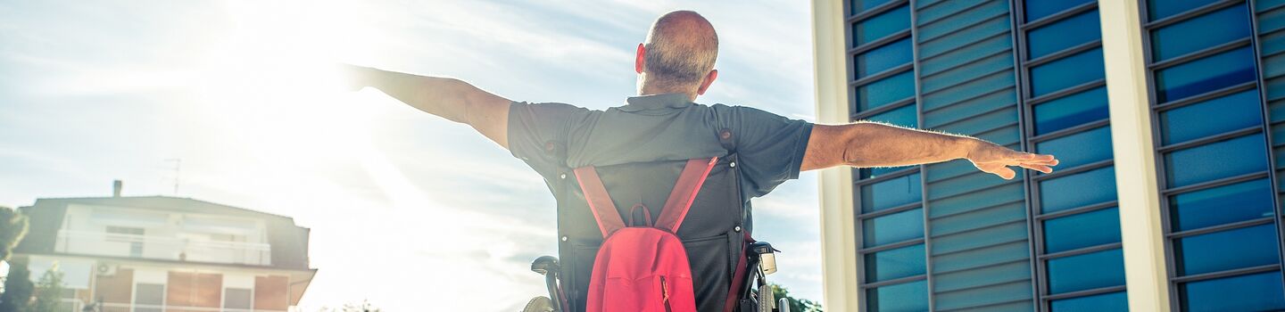 Ein Mann mit kurzen grauen Haaren sitzt in einem Rollstuhl, an der Rückseite des Rollstuhls hängt ein roter Rucksack. Der Mann hat die Arme links und rechts ausgebreitet und blickt in Richtung Sonne. Der Rollstuhl steht am Vorplatz von einem großen Gebäude.