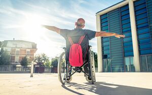 Ein Mann mit kurzen grauen Haaren sitzt in einem Rollstuhl, an der Rückseite des Rollstuhls hängt ein roter Rucksack. Der Mann hat die Arme links und rechts ausgebreitet und blickt in Richtung Sonne. Der Rollstuhl steht am Vorplatz von einem großen Gebäude.