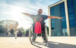 Ein Mann mit kurzen grauen Haaren sitzt in einem Rollstuhl, an der Rückseite des Rollstuhls hängt ein roter Rucksack. Der Mann hat die Arme links und rechts ausgebreitet und blickt in Richtung Sonne. Der Rollstuhl steht am Vorplatz von einem großen Gebäude.