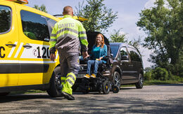 Eine Frau sitzt in einem elektrischen Rollstuhl, ein ÖAMTC Pannenfahrer in gelber Warnkleidung kommt auf sie zu. Neben den beiden steht ein gelbes ÖAMTC Pannenfahrzeug und ein schwarzer Kleinbus mit geöffneter Motorhaube.