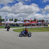 Man sieht einen Teil einer Strecke in einem Fahrtechnik Zentrum des ÖAMTC, auf der fünf Motorradfahrer unterwegs sind. Im Hintergrund sieht man Zelte diverser Motorradhersteller mit ausgestellten Fahrzeugen zum Testen.