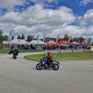 Man sieht einen Teil einer Strecke in einem Fahrtechnik Zentrum des ÖAMTC, auf der fünf Motorradfahrer unterwegs sind. Im Hintergrund sieht man Zelte diverser Motorradhersteller mit ausgestellten Fahrzeugen zum Testen.
