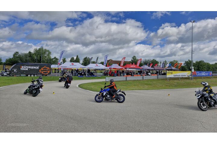 Man sieht einen Teil einer Strecke in einem Fahrtechnik Zentrum des ÖAMTC, auf der fünf Motorradfahrer unterwegs sind. Im Hintergrund sieht man Zelte diverser Motorradhersteller mit ausgestellten Fahrzeugen zum Testen.