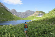 Eine Wanderin geht zu einer Hütte in den Bergen, vorbei an einem Bergsee.