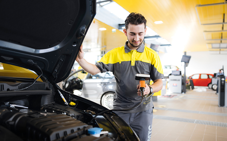 Ein Mann in grauer Arbeitskleidung kontrolliert Daten eines Fahrzeuges vor der offenen Motorhaube.