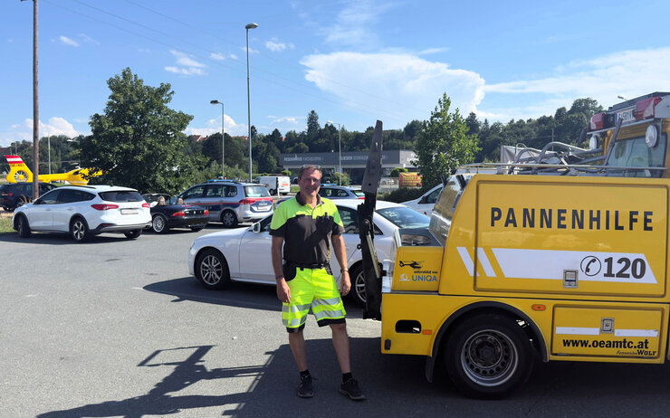 Pannenhilfe im Sommer ÖAMTC Pannenhelfer steht auf Parkplatz neben gelben Pannenhilfeauto. Im Hintergrund ist ein Christophorus-Hubschrauber zu sehen.