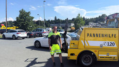 ÖAMTC Pannenhelfer steht auf Parkplatz neben gelben Pannenhilfeauto. Im Hintergrund ist ein Christophorus-Hubschrauber zu sehen.