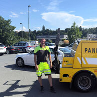 ÖAMTC Pannenhelfer steht auf Parkplatz neben gelben Pannenhilfeauto. Im Hintergrund ist ein Christophorus-Hubschrauber zu sehen.