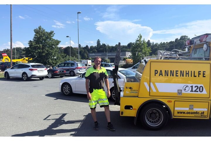 ÖAMTC Pannenhelfer steht auf Parkplatz neben gelben Pannenhilfeauto. Im Hintergrund ist ein Christophorus-Hubschrauber zu sehen.