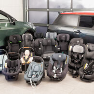 OEAMTC-Kindersitztest-Fruehjahr-2025 (1) Eine Auswahl an Kindersitzen aus dem Test steht aufgereiht auf dem Boden vor zwei Testfahrzeugen in einer Werkstatt.
