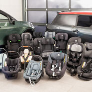 Eine Auswahl an Kindersitzen aus dem Test steht aufgereiht auf dem Boden vor zwei Testfahrzeugen in einer Werkstatt.