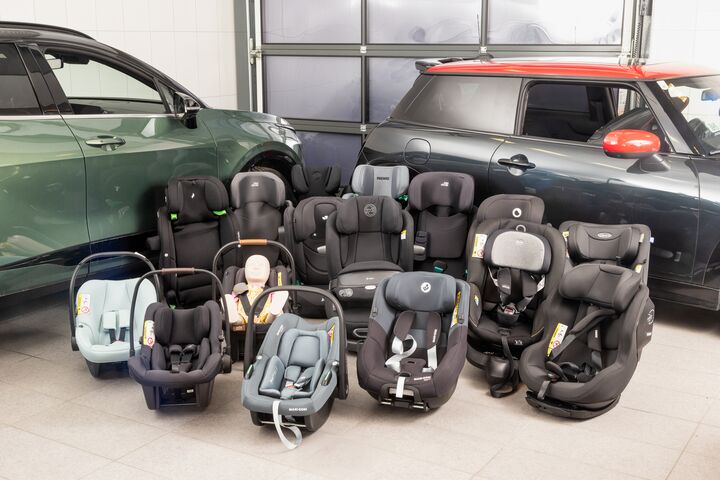 Eine Auswahl an Kindersitzen aus dem Test steht aufgereiht auf dem Boden vor zwei Testfahrzeugen in einer Werkstatt.