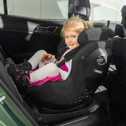 Ein kleines Mädchen mit blonden Haaren sitzt beim Kindersitztest in einem rückwärtsgerichteten Kindersitz auf der Rückbank eines Autos und lächelt.