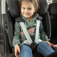 Ein kleines Mädchen mit braunen Haaren sitzt im Rahmen des Kindersitztests angeschnallt in einem Kindersitz eines Autos. Sie trägt einen grünen Pullover und blaue Jeans und lächelt.