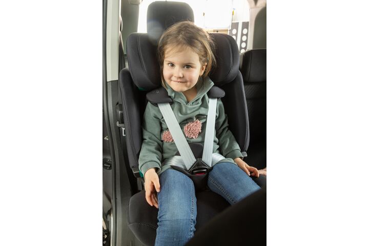 Ein kleines Mädchen mit braunen Haaren sitzt im Rahmen des Kindersitztests angeschnallt in einem Kindersitz eines Autos. Sie trägt einen grünen Pullover und blaue Jeans und lächelt.