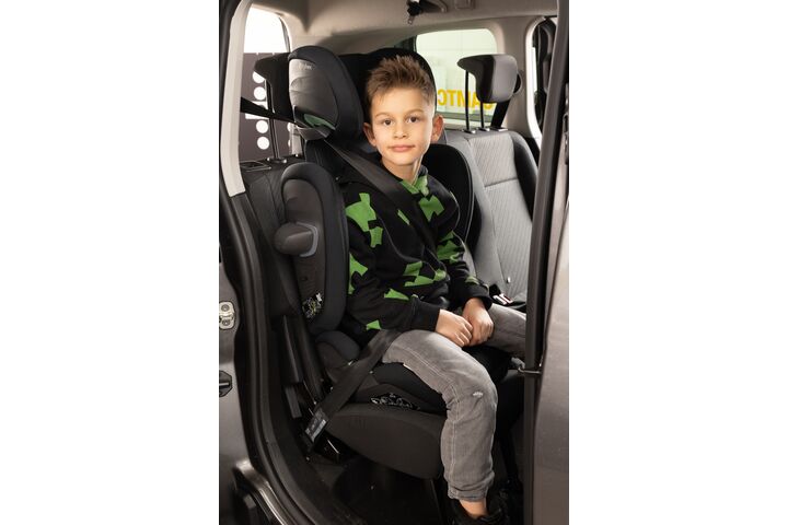 Ein Junge sitzt im Rahmen des Kindersitztests in einem Kindersitz auf der Rückbank eines Autos. Er trägt einen grün-schwarzen Pullover und eine graue Jeanshose.