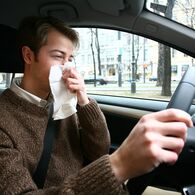 Ein junger Mann putzt sich die Nase mit einem Taschentuch, während er am Steuer eines Pkw sitzt