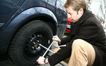 Junger Mann in Winterkleidung kniet vor Auto mit einem Radkreuzschlüssel in der Hand vor Reifen.