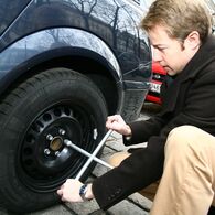 Junger Mann in Winterkleidung kniet vor Auto mit einem Radkreuzschlüssel in der Hand vor Reifen.