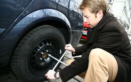Junger Mann in Winterkleidung kniet vor Auto mit einem Radkreuzschlüssel in der Hand vor Reifen.