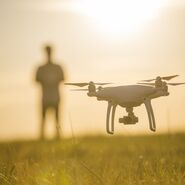 Auf dem Bild sieht man eine Drohne im Flug. Die Drohne schwebt knapp über einer Wiese. Im Hintergrund unscharf zu erkennen: Eine Person, die die Drohne steuert.