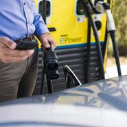 An einer ÖAMTC ePower Ladesäule steht ein Mann und hält das Ladekabel in der linken Hand, in der rechten Hand hält er ein Smartphone. Vor ihm steht ein Auto mit offener Ladebuchse.