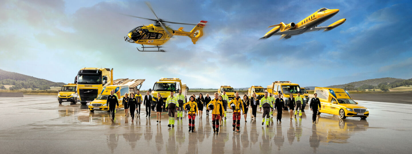 Eine Flotte von gelben Rettungsfahrzeugen und Luftfahrzeugen steht auf einem Flugfeld. Zu sehen ein Krankenwagen, Pannenfahrzeuge, ein Abschlepp-LKW, ein Rettungshubschrauber in der Luft sowie ein Ambulanzflugzeug. Davor stehen Einsatzkräfte in leuchtender Schutzkleidung. 