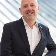 Portrait eines Mannes der am Geländer lehnt, einen weißes Hemd und einen schwarze Anzugjacke trägt.