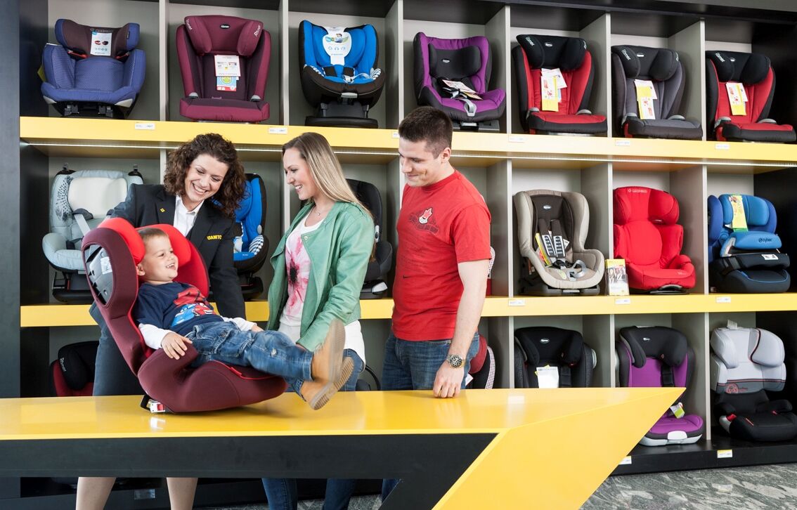 An einem ÖAMTC Standort sind in einem großen Wandregal viele verschiedene Kindersitze ausgestellt. Im Vordergrund ist ein Tisch, dahinter stehen eine ÖAMTC Mitarbeiterin, eine weitere Frau und ein Mann. Am Tisch steht ein roter Kindersitz und darin sitzt ein kleiner Bub, dieser probiert fröhlich den neuen Kindersitz.