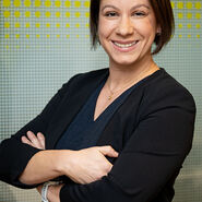 Porträtfoto einer Frau mit mittellangen braunen Haaren, die lächelnd und mit verschränkten Armen vor einer gelben-weißen Glaswand steht