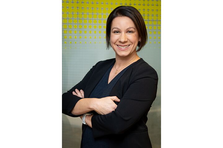 Porträtfoto einer Frau mit mittellangen braunen Haaren, die lächelnd und mit verschränkten Armen vor einer gelben-weißen Glaswand steht