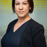 Porträtfoto einer Frau mit mittellangen braunen Haaren, die vor einer gelben-weißen Glaswand steht