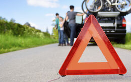 Ein pannendreieck steht auf einer Straße. Im Hintergrund ist man ein schwarzes Auto, das steht, mit Fahrrädern am Dach und eine Familie, die neben dem Auto steht.
