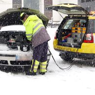 EIn Pannenhelfer steht vor einem Auto mit geöffneter Motorhaube und gibt Starthilfe. Schnee liegt auf der Straße und es schneit.