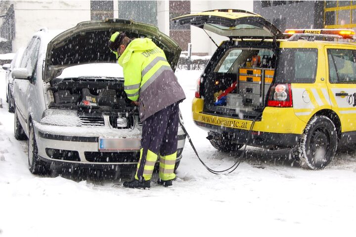 EIn Pannenhelfer steht vor einem Auto mit geöffneter Motorhaube und gibt Starthilfe. Schnee liegt auf der Straße und es schneit.