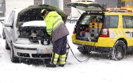 EIn Pannenhelfer steht vor einem Auto mit geöffneter Motorhaube und gibt Starthilfe. Schnee liegt auf der Straße und es schneit.