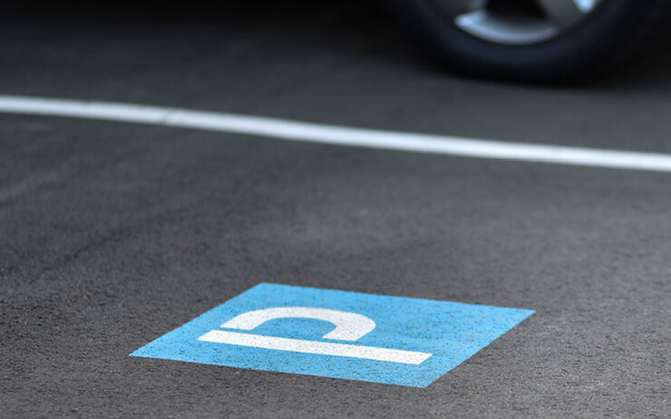 Parken - Parkplatz Kennzeichnung eines Parkplatzes mit dem Symbol P am Boden (weiß auf blauem Hintergrund) und weißer Randlinie; hinter der weißen Linie sieht man ein geparktes Auto.