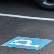 Kennzeichnung eines Parkplatzes mit dem Symbol P am Boden (weiß auf blauem Hintergrund) und weißer Randlinie; hinter der weißen Linie sieht man ein geparktes Auto.