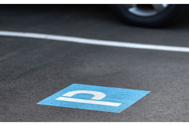 Kennzeichnung eines Parkplatzes mit dem Symbol P am Boden (weiß auf blauem Hintergrund) und weißer Randlinie; hinter der weißen Linie sieht man ein geparktes Auto.