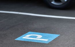 Kennzeichnung eines Parkplatzes mit dem Symbol P am Boden (weiß auf blauem Hintergrund) und weißer Randlinie; hinter der weißen Linie sieht man ein geparktes Auto.