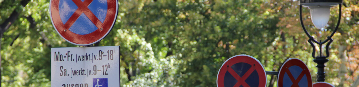 Mehrer Halten & Parken verboten-Verkehrsschilder mit Zusatztafeln, die auf einer Straße mit Bäumen hintereinander aufgestellt sind.