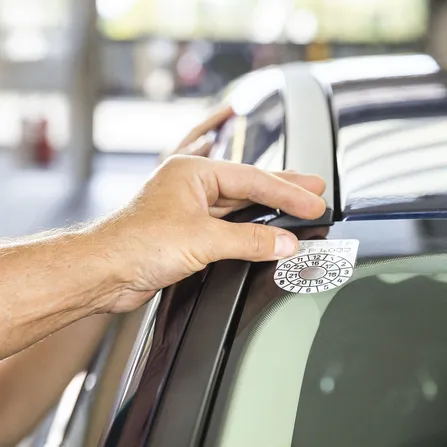 Hand von ÖAMTC-Techniker klebt §57a-Plakette - Pickerl - auf das Auto. Im Hintergrund steht eine Frau.