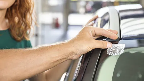 Hand von ÖAMTC-Techniker klebt §57a-Plakette - Pickerl - auf das Auto. Im Hintergrund steht eine Frau.