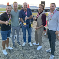 Im Bild zu sehen sind die Erstplatzierten des Team Haas bei der Siegerehrung, sie tragen eine Medaille um den Hals und halten einen Pokal und eine Flasche in die Kamera