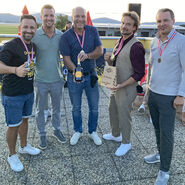 Im Bild zu sehen sind die Erstplatzierten des Team Haas bei der Siegerehrung, sie tragen eine Medaille um den Hals und halten einen Pokal und eine Flasche in die Kamera