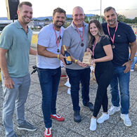 Im Bild zu sehen sind die Drittplatzierten des Team Orehounig bei der Siegerehrung, sie tragen eine Medaille um den Hals und halten einen Pokal und eine Flasche in die Kamera