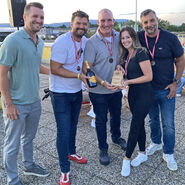 Im Bild zu sehen sind die Drittplatzierten des Team Orehounig bei der Siegerehrung, sie tragen eine Medaille um den Hals und halten einen Pokal und eine Flasche in die Kamera